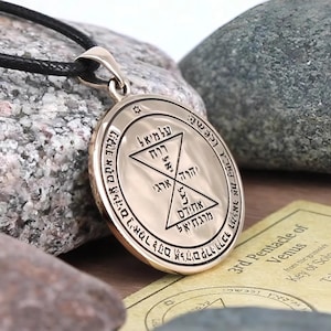 May include: Silver pendant with a black cord. The pendant features an engraved design with Hebrew text and geometric shapes. The text includes the words "3rd Pentacle of Venus" and "Key of Solomon". The pendant is displayed on a stone surface.