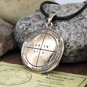 May include: A gold-colored pendant with Hebrew text and symbols, suspended from a black cord. The pendant features a circular design with intricate details and is accompanied by a paper with additional text. The text on the paper reads "5th Pentacle of Mercury Key of Solomon".