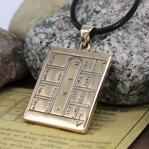 May include: A gold-colored rectangular pendant with etched symbols and text, suspended from a black cord. The pendant has a detailed design with various shapes and lines. The background includes a rock and a piece of paper with text.