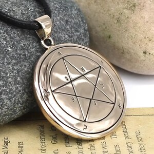 May include: A silver-colored pendant with a black cord. The pendant features a seven-pointed star within a circle, with symbols and text around the edges. The pendant is likely made of metal and is displayed against a stone background.