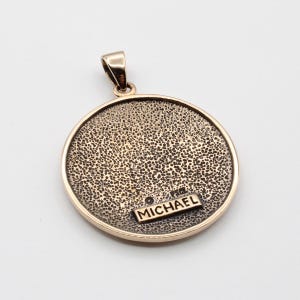 May include: A gold-toned pendant with a textured surface and a small rectangular tag that reads "MICHAEL". The pendant has a circular shape with a loop for a chain. The overall design is simple and elegant.