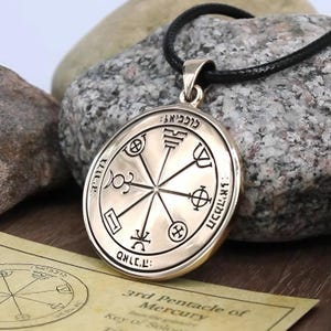 May include: A gold-colored pendant with intricate symbols and Hebrew text, suspended on a black cord. The pendant features a circular design with various shapes and lines. A paper with text is visible in the foreground.