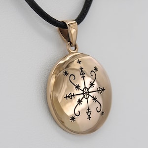 May include: A gold-colored pendant with a black cord. The pendant is round with a detailed black design in the center. The design includes a central star-like shape with arrows and other symbols. The pendant is suspended from a black cord.