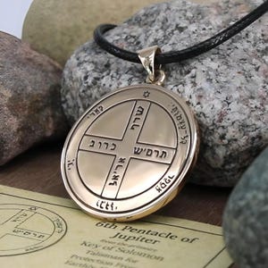 May include: A gold-colored pendant with a black cord. The pendant features engraved Hebrew text and symbols within a circular design. The text "6th Pentacle of Jupiter" is visible on a paper below the pendant. The pendant is displayed on a stone surface.