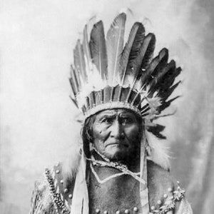 May include: A black and white portrait of a Native American man wearing a traditional headdress with feathers. He is looking directly at the camera with a serious expression.