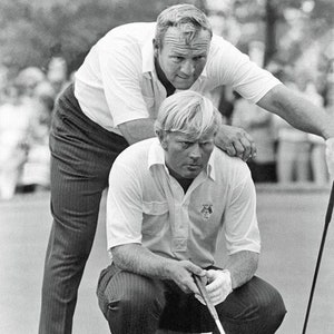 May include: Two men on a golf course, one is kneeling and holding a golf club, the other is standing behind him with his arm around his shoulder.