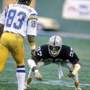 May include: Two football players in vintage uniforms on a green field. One player wears a white and blue jersey with the number 83 and the name "Jefferson". The other player is in a black and silver uniform, in a defensive stance.