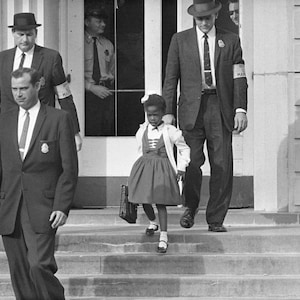 May include: A young girl in a dress and cardigan walks down a set of stairs, escorted by two men in suits. The men are wearing badges and the girl is carrying a book bag.