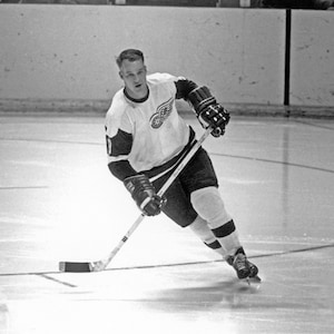 May include: A black and white photo of a hockey player wearing a white jersey with a red winged logo. The player is skating on the ice and holding a hockey stick.