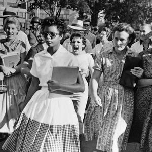 May include: A black and white photo of a group of people, mostly women, walking down a street. Some of the women are holding books and others are holding purses. The women are dressed in dresses and skirts. The photo appears to be from the 1950s or 1960s.