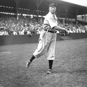May include: A black and white photo of a baseball player in a uniform, pitching on a baseball field. The player is wearing a cap with a letter "G" on it.