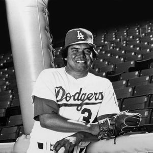 May include: A baseball player wearing a white Dodgers jersey with the number 2 on the front, a blue and white LA baseball cap, and a black and brown baseball glove.