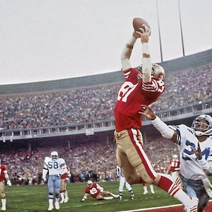 May include: A football player in a red and gold uniform jumps to catch a football thrown by a player in a white and blue uniform. The game is being played in a stadium with a large crowd of spectators.