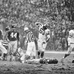 May include: Black and white photo of a football game in progress. Players in uniform are running and tackling on the field. A referee is standing in the middle of the action.