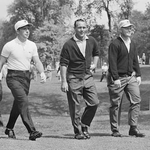 May include: Three men wearing hats and casual clothing walk on a grassy field. The man in the middle is wearing a dark cardigan sweater and light-colored pants. The man on the left is wearing a white polo shirt and dark pants. The man on the right is wearing a dark sweater and light-colored pants.