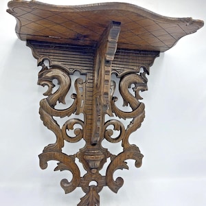 May include: Ornate, carved wooden wall shelf. The shelf has a curved top with a diamond pattern and is supported by intricately carved brackets featuring scrollwork and stylized animal heads. The wood has a warm, brown tone.