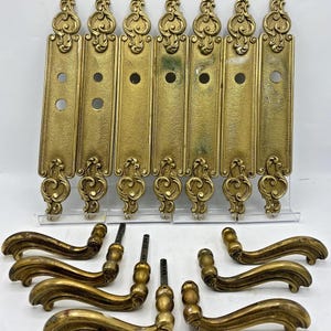 Puede incluir: Un conjunto de siete placas de puerta rectangulares de latón con volutas ornamentales en la parte superior e inferior, con aberturas circulares. Incluye seis manijas de puerta curvas de latón con acabado metálico y signos de desgaste.