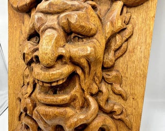 May include: A detailed, hand-carved wooden wall sculpture depicting a grinning, horned face with a prominent nose and beard. The sculpture is made of light brown wood, with visible grain and texture, and is mounted on a rectangular wooden panel.