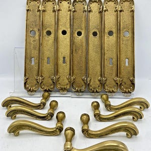 May include: A set of seven rectangular brass door plates with ornate scrollwork at the top and bottom, each featuring a circular knob hole and a keyhole. Seven matching curved brass door handles are arranged in front of the plates.