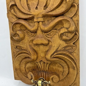 May include: A carved wooden wall plaque depicting a stylized face with intricate details. The plaque is a warm brown colour and has a brass door knocker attached at the bottom. The design is reminiscent of classical architectural elements.