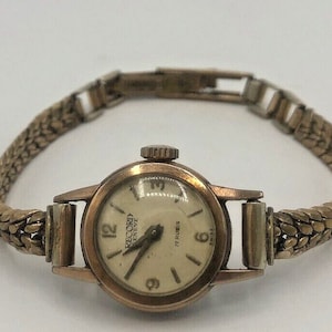 Op de afbeelding: Een gouden polshorloge met een witte wijzerplaat en zwarte wijzers. De wijzerplaat van het horloge heeft zwarte Romeinse cijfers als uurmarkeringen en de tekst "RECORD GENEVE 17 RUBIS" erop gedrukt. Het horloge heeft een gouden metalen band.