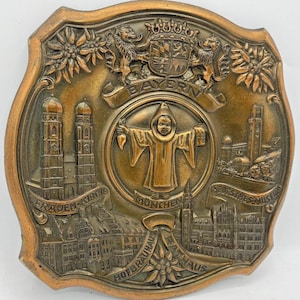 Pode incluir: Placa decorativa em bronze com motivos bávaros. Inclui representações de Frauenkirche, Deutsches Museum e Hofbräuhaus. A placa tem uma figura central com uma túnica com capuz, rodeada por texto e detalhes florais.