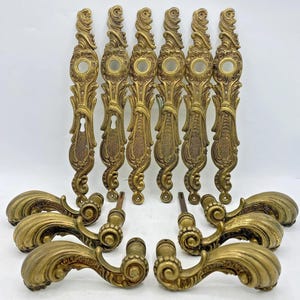 Peut inclure: Ensemble de quincaillerie de porte ancienne en laiton, comprenant des plaques arrière ornées avec des trous de serrure et des accents circulaires, ainsi que des poignées de porte incurvées assorties. La quincaillerie présente des volutes détaillées et une patine chaude vieillie, suggérant un style vintage.