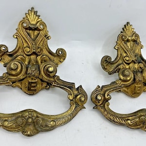 Puede incluir: Dos tiradores de cajón antiguos de latón dorado con intrincados adornos y un emblema "E". Estos tiradores dorados tienen un diseño de asa curvada, ideales para la restauración de muebles antiguos. Miden unos 15 cm de largo.