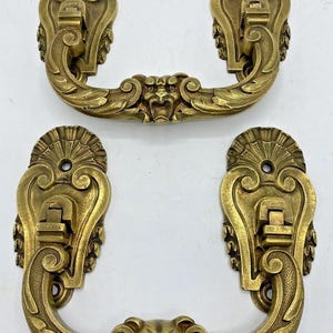 Puede incluir: Dos tiradores de cajón de latón a juego con un diseño ornamentado y simétrico con un motivo central de cabeza de león. Cada tirador tiene dos placas de montaje con partes superiores en forma de abanico y brazos curvos.