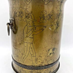 Könnte beinhalten: Ein Messing-Pflanztopf mit einem gravierten Design eines Jägers mit einer Waffe, einem Hund und einem Vogel. Der Topf hat ein dekoratives Band um den oberen und unteren Rand und steht auf Holzfüßen.