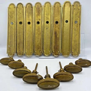 Peut inclure: Un ensemble de huit plaques de porte rectangulaires en laiton avec des extrémités arrondies et un trou circulaire central, accompagné de huit poignées de porte ovales en laiton assorties avec des bords perlés et des tiges métalliques.
