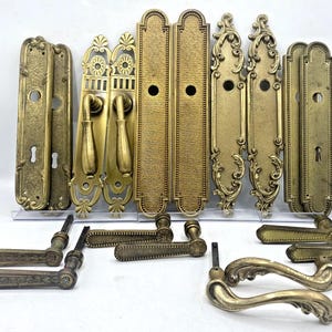 Peut inclure: Une collection de quincaillerie de porte en laiton comprenant plusieurs plaques arrière rectangulaires avec des trous de serrure et des ouvertures circulaires, ainsi que diverses poignées de porte assorties. Les pièces présentent des volutes ornées.