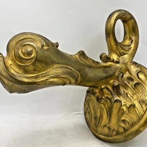 Könnte beinhalten: Verziertes, goldfarbenes dekoratives Architekturelement mit einem wirbelnden, blattartigen Design. Das Stück hat einen kreisförmigen Sockel und eine gebogene, offene Schlaufe an der Oberseite. Die Oberfläche hat ein strukturiertes, gealtertes Aussehen.