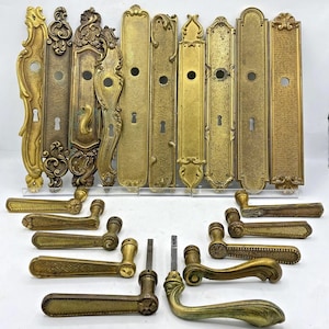 Peut inclure: Une collection de quincaillerie de porte en laiton antique comprenant huit plaques de propreté rectangulaires et ornées avec trous de serrure, ainsi que huit poignées de porte en laiton assorties aux finitions métalliques variées.