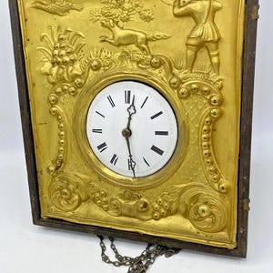 Op de afbeelding: Antieke goudkleurige wandklok met een jachtscène reliëf. De wijzerplaat is wit met zwarte Romeinse cijfers. De klok heeft een donkerbruin houten frame en een decoratieve ketting.