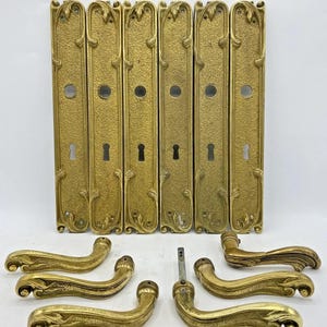 Peut inclure: Ensemble de quincaillerie de porte ancienne en laiton, comprenant six plaques arrière rectangulaires avec trous de serrure et trous ronds, et six poignées de porte incurvées. La quincaillerie présente un design orné et texturé avec des motifs floraux et de feuilles.