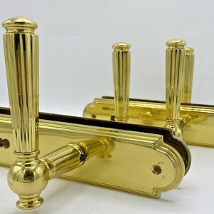 Peut inclure: Deux ensembles de poignées de porte dorées. Chaque poignée a une longue poignée cannelée et une base arrondie. Les plaques arrière sont rectangulaires avec des bords arrondis. Les poignées sont en métal brillant et réfléchissant.