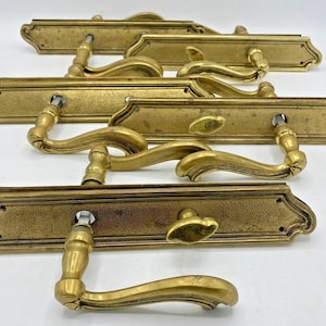 Op de afbeelding: Een verzameling antieke messing deurklinken. Elke klink heeft een decoratieve achterplaat met een getextureerd oppervlak en een gebogen hendel. De klinken zijn goudkleurig en lijken vintage te zijn.