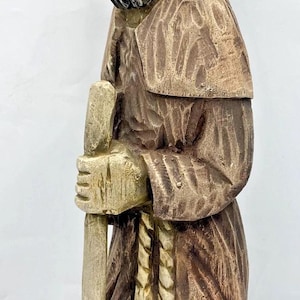 Pode incluir: Estátua de madeira esculpida de um homem barbudo em uma túnica com capuz, segurando um bastão. A estátua é principalmente marrom com detalhes mais claros, mostrando entalhes detalhados e uma estética rústica. A figura parece ser um personagem religioso ou histórico.