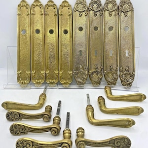 May include: A collection of eight brass door plates and eight matching brass door handles. Four plates feature a central circular hole, while four include a circular hole and a keyhole. All pieces display ornate, embossed scrollwork detailing.