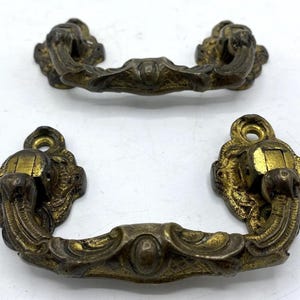 Puede incluir: Dos tiradores de cajón antiguos de bronce con detalles ornamentados. Cada tirador presenta un asa curvada con un diseño ovalado central y piezas finales decorativas. El herraje tiene un aspecto vintage y envejecido con toques dorados.