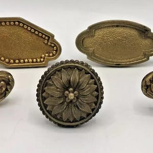 Puede incluir: Cinco pomos de armario de latón antiguo con diseños ornamentados. Los pomos presentan una variedad de patrones, incluyendo florales, geométricos y de volutas.
