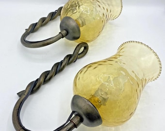 Op de afbeelding: Twee vintage wandkandelaars met amberkleurige glazen kappen en gedraaide metalen armen. De kappen hebben een getextureerd oppervlak en een decoratieve rand. De metalen armen zijn donkerbruin met een lusontwerp om op te hangen.