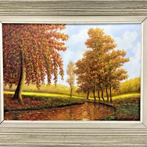 Könnte beinhalten: Ölgemälde einer Herbstlandschaft mit Bäumen in Orange-, Rot- und Gelbtönen, die sich in einem ruhigen Fluss spiegeln. Das Kunstwerk ist in einem weißen, verwitterten Holzrahmen gerahmt, was eine rustikale Ästhetik erzeugt.