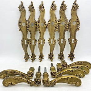 Op de afbeelding: Een verzameling antieke messing deurbeslagen, waaronder sierlijke achterplaten, deurgrepen en sleutelgat rozetten. De hardware heeft een gedetailleerd, opgerold ontwerp en een warme, verouderde patina. De set bevat meerdere onderdelen.