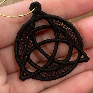 May include: Black lace earring featuring a Celtic knot design. The intricate openwork pattern is suspended from a gold-colored hoop. The earring is held in a hand, showcasing the delicate craftsmanship.