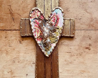 May include: Wooden cross with a heart-shaped centerpiece. The heart features a floral design with an angel figure. The cross has a gold-painted border and is set against a wooden background.