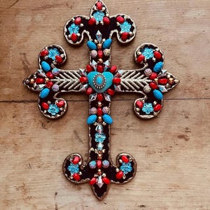May include: Ornate, handcrafted cross with a black base, adorned with turquoise, red, and clear gemstones. The cross features gold-colored filigree and a turquoise heart-shaped center. Decorative religious item.