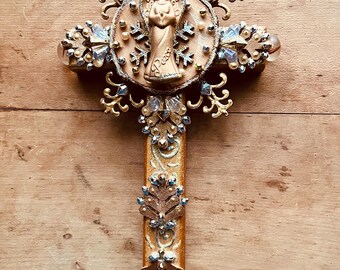 May include: Ornate gold-colored cross with an angel figure at the center. The cross features intricate detailing, including clear and blue crystal accents, and decorative flourishes. The word "Peace" is inscribed on the cross.