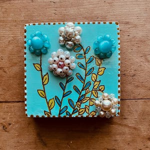 May include: A square, hand-painted wooden plaque with a turquoise background. The plaque features four beaded flower embellishments in shades of white, pink, and blue. Gold and blue painted stems and leaves complete the floral design.